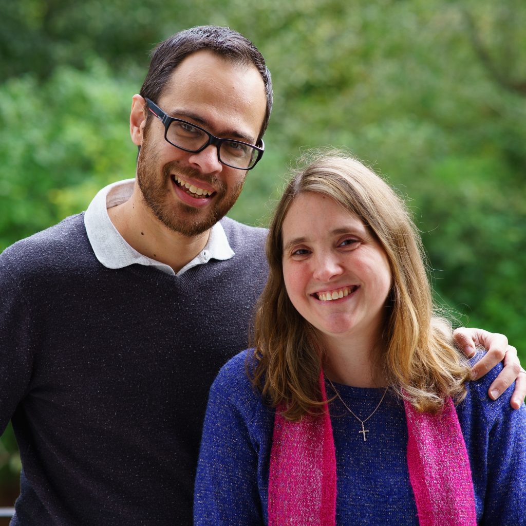 Photo of Dan Acharya (a mixed race man with glasses and a beard) and Jo Acharya (a white woman with long brown hair and a pink scarf), both smiling to the camera