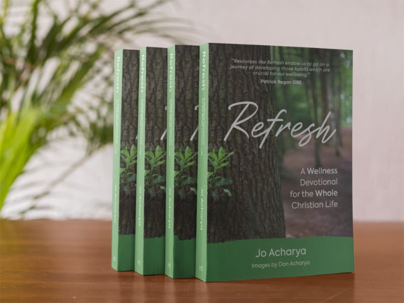 Four copies of Jo Acharya's book 'Refresh: a wellness devotional for the whole Christian life' lined up on a shelf with a leafy fern