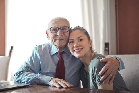 An elderly man and young woman hugging in a living room with smiles on their faces