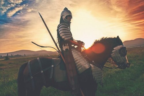 An armoured knight on a brown horse with a bright sunset behind