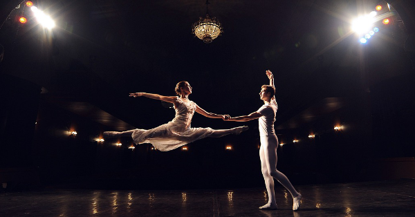 A female ballet dancer poses in the air with arms outstretched, mid-leap, holding the hand of her partner who is ready to catch her