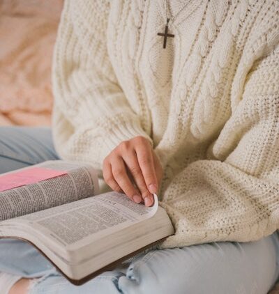 A white woman wearing a white knit jumper and a cross necklace turns the pages of the Bible, learning how the Bible helps us in our lives