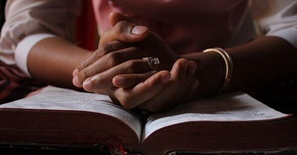 A black woman clasps her hands in prayer over an open Bible