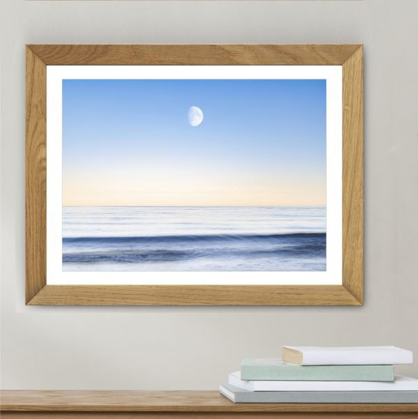 The sky and the sea print framed on a wall with a shelf and books in the foreground